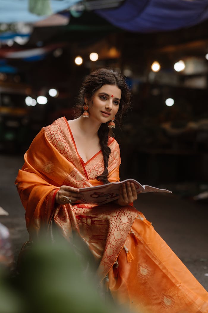 Beautiful woman in an orange saree reading a magazine at night market.