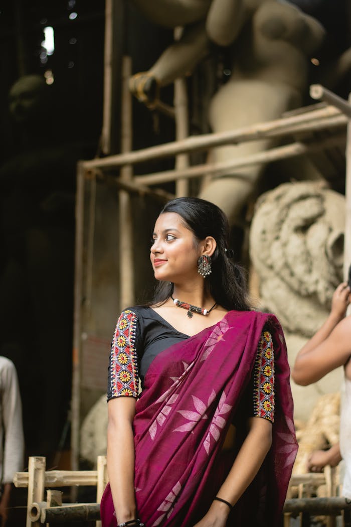 Elegant woman in traditional sari standing amidst sculptures in a dimly-lit artisan workshop.